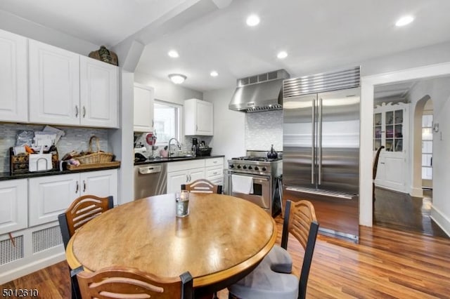 kitchen featuring high end appliances, wall chimney exhaust hood, dark countertops, backsplash, and recessed lighting