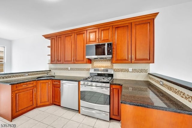 kitchen featuring appliances with stainless steel finishes, a peninsula, backsplash, dark stone countertops, and brown cabinetry