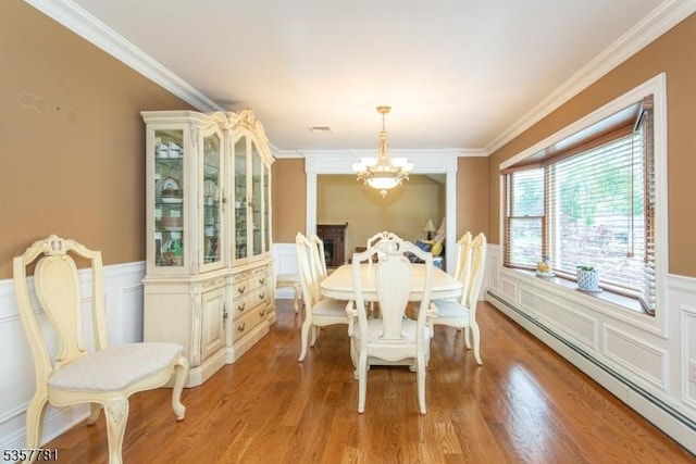 dining space featuring baseboard heating, a chandelier, a decorative wall, wainscoting, and crown molding