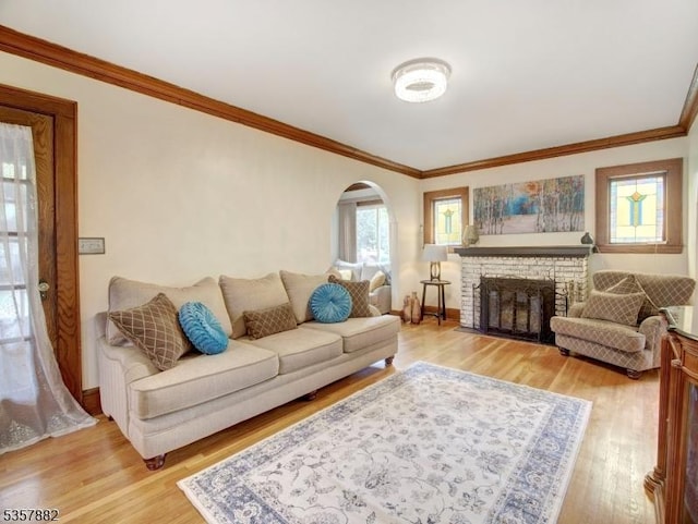 living room featuring arched walkways, light wood finished floors, ornamental molding, a brick fireplace, and baseboards