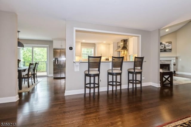 kitchen featuring stainless steel refrigerator with ice dispenser, wall chimney exhaust hood, dark wood-style floors, a kitchen breakfast bar, and white cabinetry