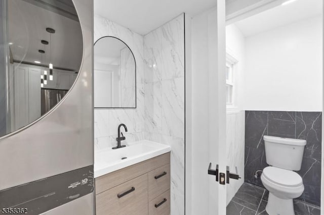 bathroom with tile walls, marble look tiles, toilet, vanity, and a wainscoted wall