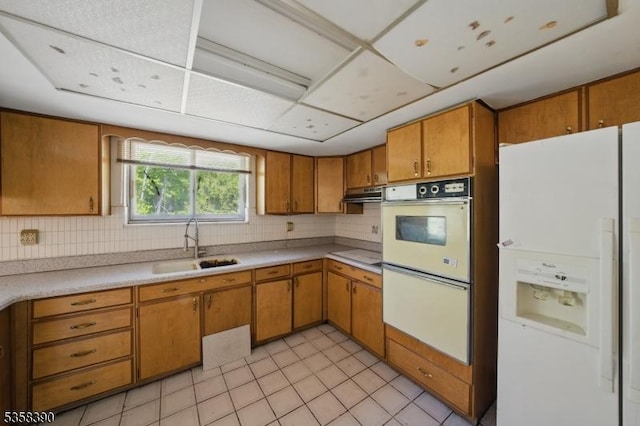 kitchen with white appliances, a sink, brown cabinets, under cabinet range hood, and tasteful backsplash