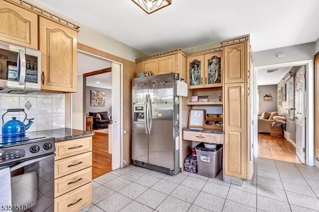kitchen featuring appliances with stainless steel finishes, light brown cabinetry, decorative backsplash, dark stone countertops, and glass insert cabinets