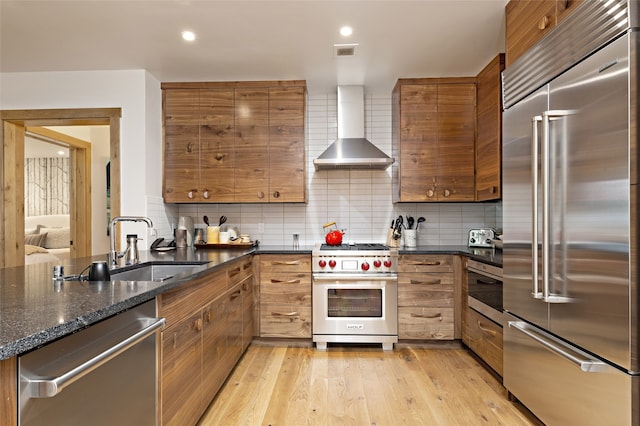 kitchen featuring wall chimney range hood, light wood-type flooring, high end appliances, backsplash, and dark stone countertops