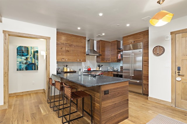 kitchen with backsplash, a kitchen breakfast bar, light hardwood / wood-style floors, stainless steel built in refrigerator, and wall chimney exhaust hood