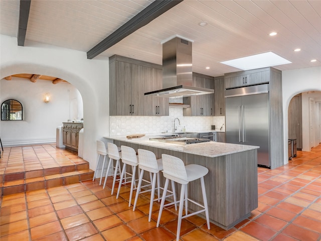 kitchen featuring tasteful backsplash, light tile flooring, island range hood, stainless steel built in refrigerator, and beam ceiling