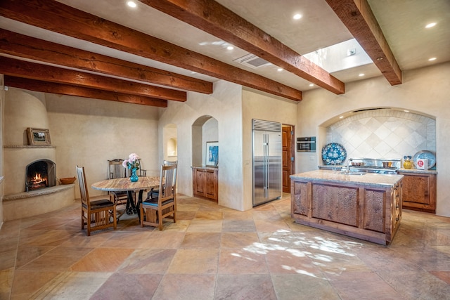 kitchen featuring built in refrigerator, a kitchen island with sink, backsplash, a fireplace, and light stone countertops