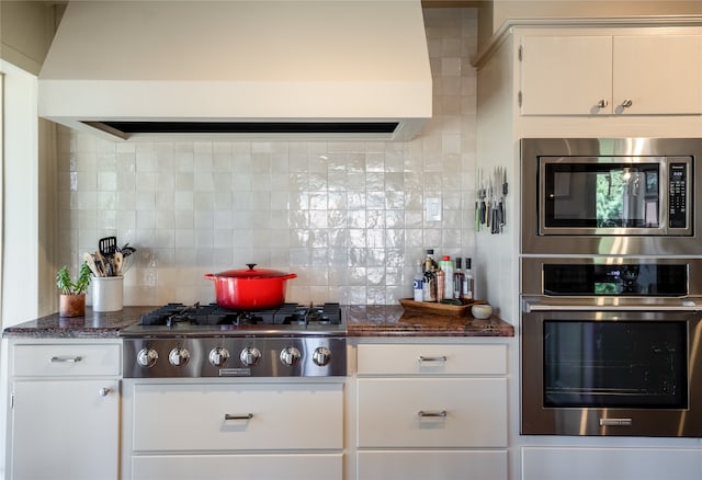 kitchen featuring white cabinetry, backsplash, appliances with stainless steel finishes, premium range hood, and dark stone countertops