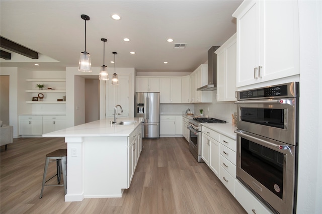 kitchen with stainless steel appliances, wall chimney range hood, white cabinetry, a kitchen island with sink, and sink