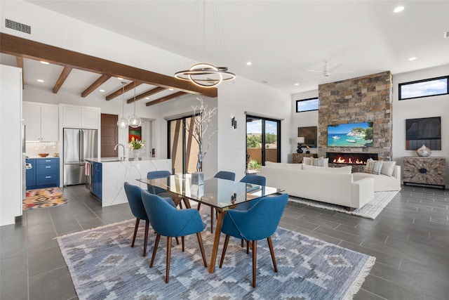 tiled dining area with ceiling fan with notable chandelier, beam ceiling, sink, and a stone fireplace