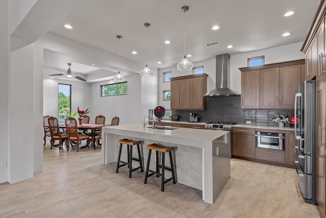 kitchen with decorative light fixtures, ceiling fan, a kitchen island with sink, stainless steel appliances, and wall chimney exhaust hood