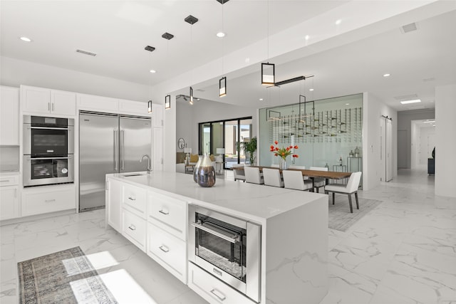 kitchen with light stone counters, a kitchen island with sink, pendant lighting, light tile patterned floors, and stainless steel appliances