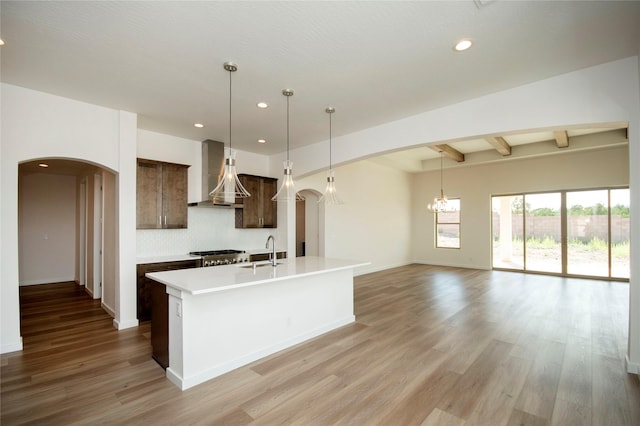 kitchen with dark brown cabinetry, sink, wall chimney range hood, a kitchen island with sink, and light wood-type flooring