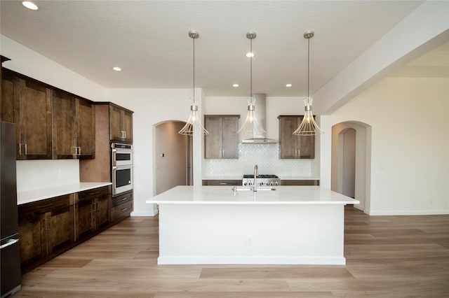 kitchen with a kitchen island with sink, light hardwood / wood-style flooring, wall chimney exhaust hood, appliances with stainless steel finishes, and dark brown cabinetry