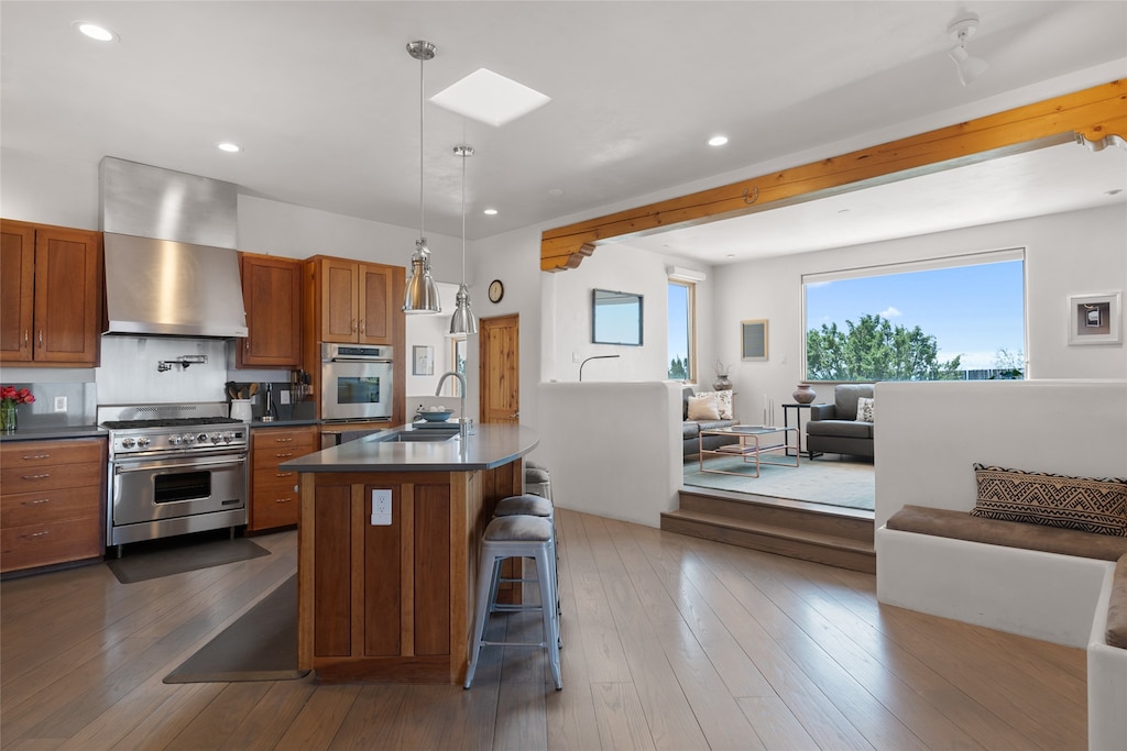 kitchen featuring stainless steel appliances, dark countertops, open floor plan, a sink, and wall chimney exhaust hood