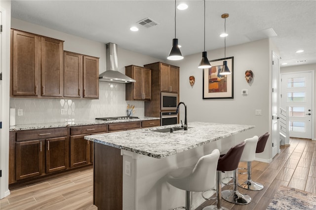 kitchen with a kitchen island with sink, wall chimney range hood, sink, hanging light fixtures, and light wood-type flooring