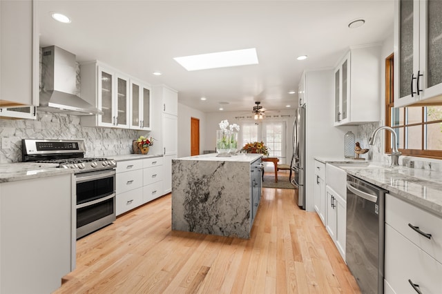 kitchen featuring a skylight, stainless steel appliances, white cabinets, and wall chimney range hood