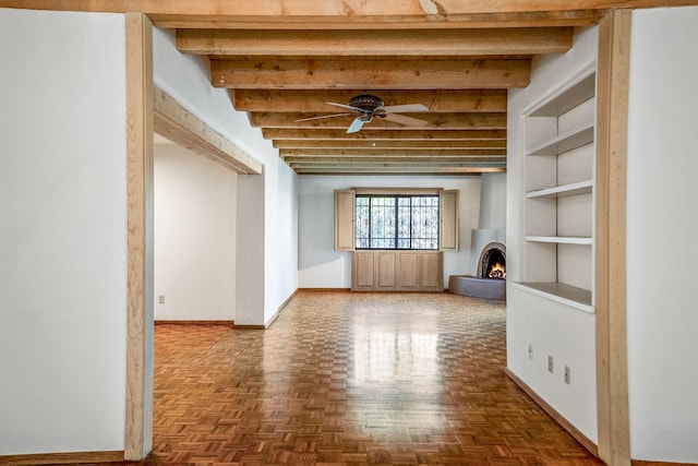 empty room with a ceiling fan, a lit fireplace, baseboards, and beam ceiling