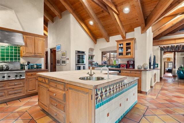 kitchen featuring appliances with stainless steel finishes, wall chimney range hood, sink, beamed ceiling, and an island with sink