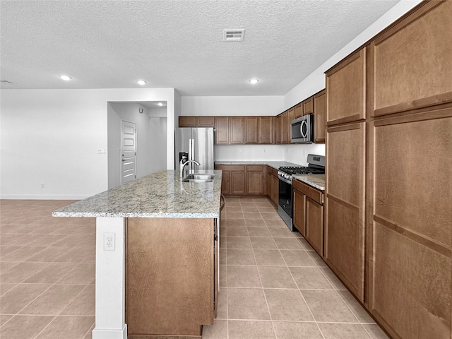 kitchen featuring a kitchen island with sink, light stone countertops, a textured ceiling, light tile patterned floors, and appliances with stainless steel finishes