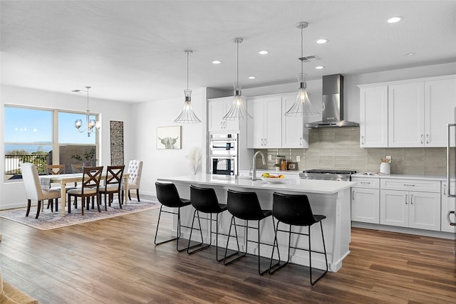 kitchen featuring hanging light fixtures, double oven, wall chimney range hood, a kitchen island with sink, and white cabinets