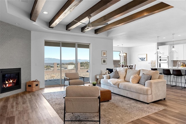 living room with ceiling fan with notable chandelier, a large fireplace, light hardwood / wood-style floors, a mountain view, and beam ceiling