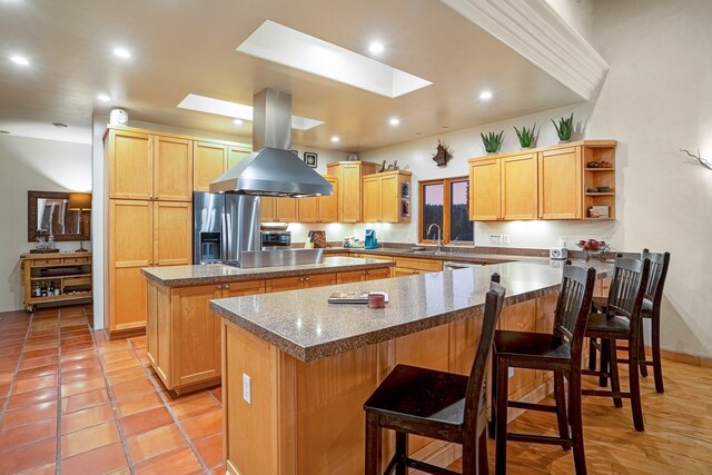 kitchen featuring a skylight, stainless steel refrigerator with ice dispenser, island range hood, a kitchen bar, and a kitchen island