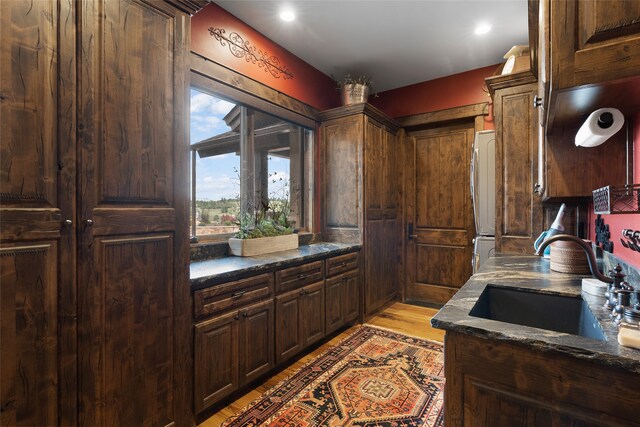 kitchen featuring dark stone counters, light hardwood / wood-style flooring, dark brown cabinetry, sink, and stainless steel fridge