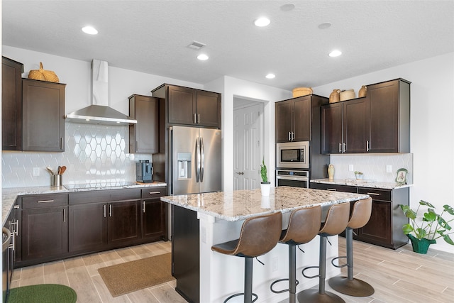 kitchen with appliances with stainless steel finishes, a kitchen bar, a center island, decorative backsplash, and wall chimney range hood