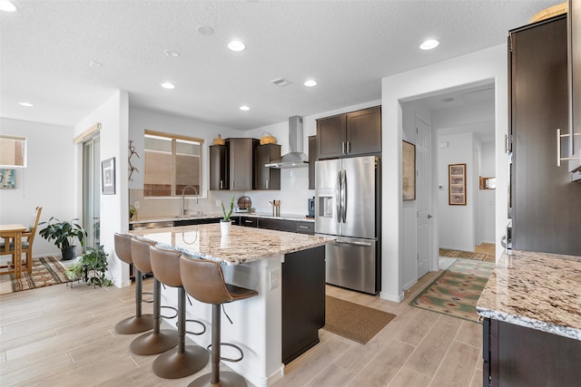 kitchen featuring stainless steel fridge, light stone counters, a center island, a breakfast bar, and wall chimney exhaust hood