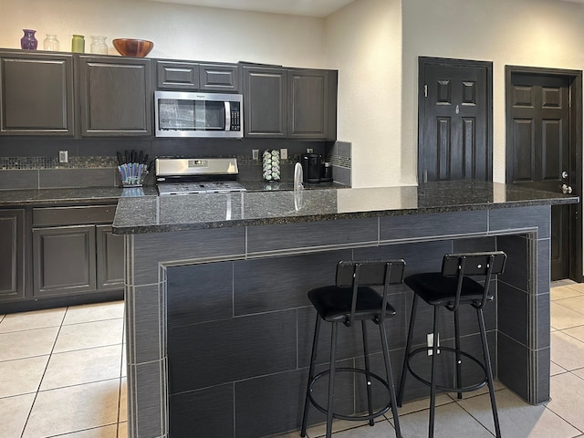 kitchen with a breakfast bar, appliances with stainless steel finishes, light tile patterned floors, and dark stone counters