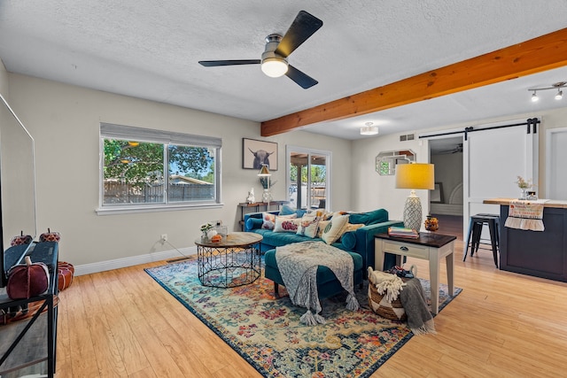 living room with a textured ceiling, beam ceiling, light hardwood / wood-style floors, a barn door, and ceiling fan