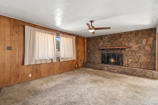 unfurnished living room featuring ceiling fan, wood walls, a stone fireplace, a textured ceiling, and carpet