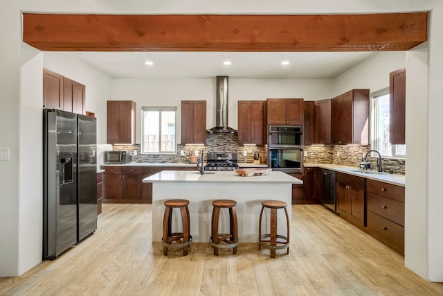 kitchen with wall chimney range hood, a kitchen island with sink, a kitchen bar, light wood-type flooring, and stainless steel appliances