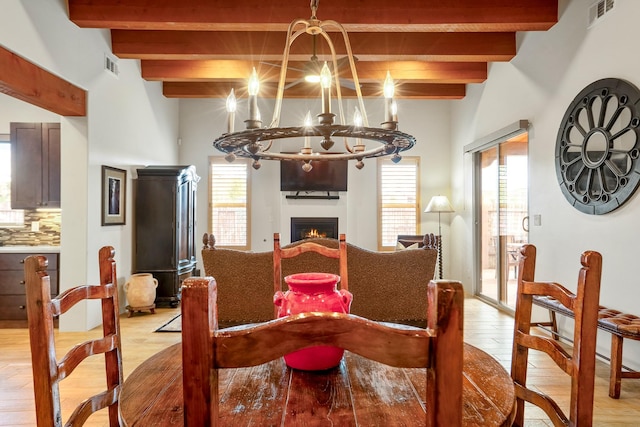 dining area with a chandelier, a warm lit fireplace, light wood-style flooring, visible vents, and beam ceiling