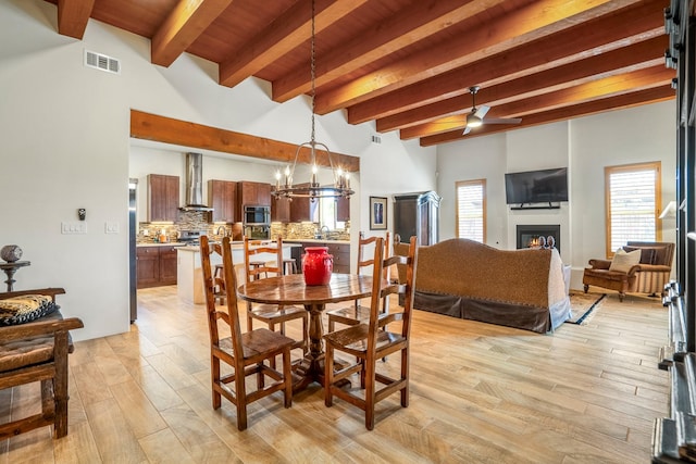 dining area with a warm lit fireplace, ceiling fan with notable chandelier, visible vents, light wood-style floors, and beam ceiling