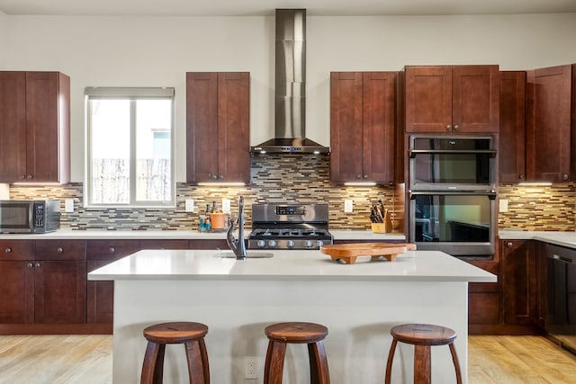 kitchen featuring wall chimney range hood, a kitchen breakfast bar, light hardwood / wood-style flooring, stainless steel appliances, and a center island with sink