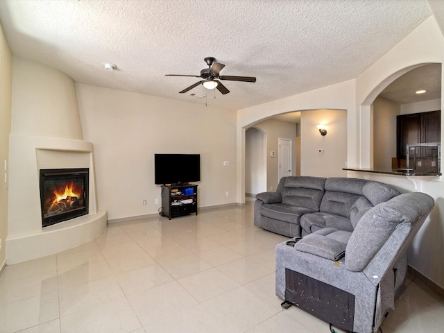 living room featuring arched walkways, a large fireplace, ceiling fan, a textured ceiling, and baseboards