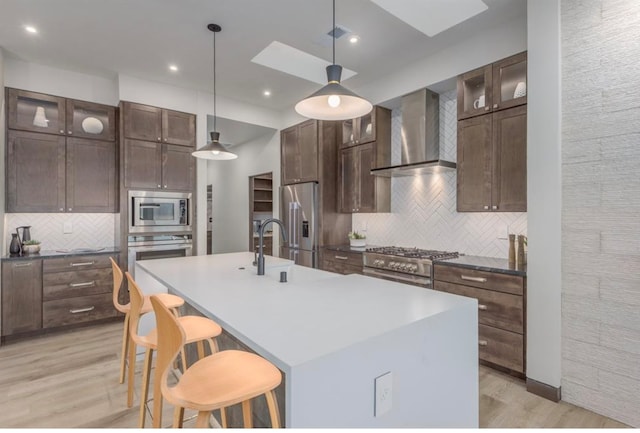 kitchen with decorative light fixtures, a kitchen island with sink, wall chimney range hood, appliances with stainless steel finishes, and a kitchen breakfast bar