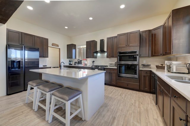 kitchen with wall chimney range hood, an island with sink, light wood-type flooring, a kitchen bar, and stainless steel appliances