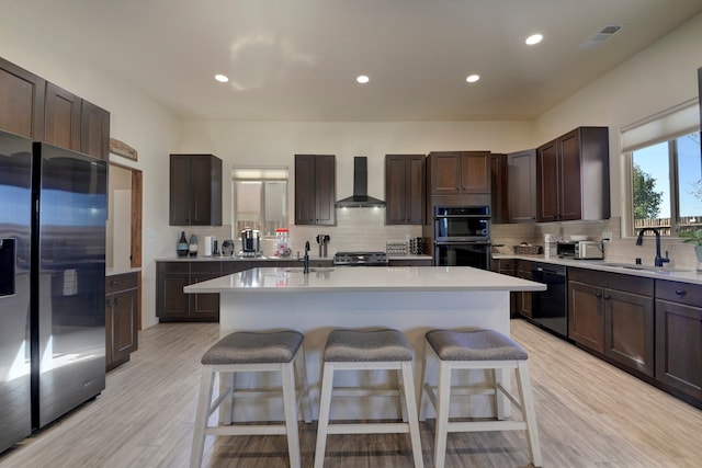 kitchen with wall chimney range hood, an island with sink, stainless steel appliances, sink, and light hardwood / wood-style floors