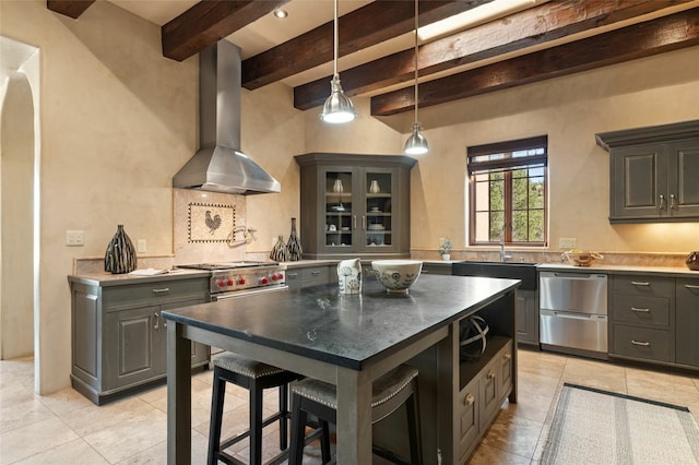 kitchen with wall chimney range hood, appliances with stainless steel finishes, a kitchen island, beam ceiling, and light tile patterned floors