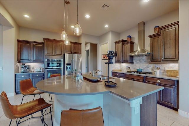 kitchen featuring a center island with sink, sink, wall chimney exhaust hood, appliances with stainless steel finishes, and decorative light fixtures