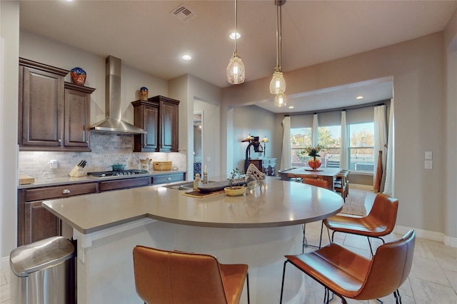 kitchen with wall chimney exhaust hood, a kitchen breakfast bar, an island with sink, pendant lighting, and stainless steel gas stovetop