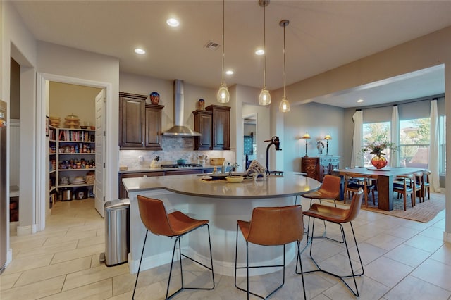 kitchen featuring a breakfast bar, decorative light fixtures, a center island with sink, and wall chimney exhaust hood