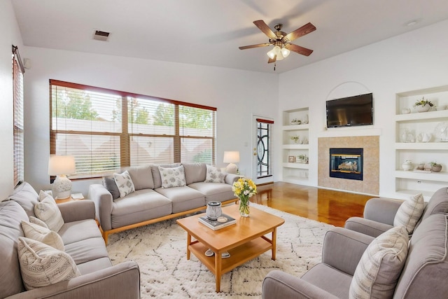 living room featuring a tile fireplace, vaulted ceiling, ceiling fan, built in features, and light wood-type flooring