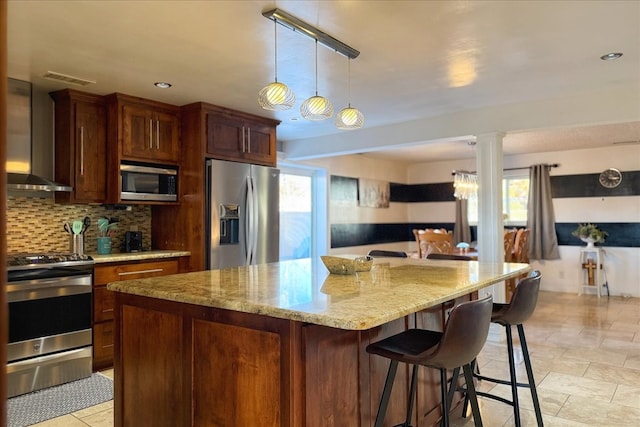 kitchen featuring hanging light fixtures, wall chimney exhaust hood, a kitchen island, light stone counters, and stainless steel appliances