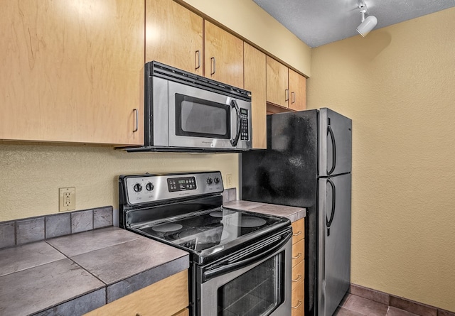 kitchen with light brown cabinets, stainless steel appliances, dark tile patterned floors, track lighting, and a textured ceiling