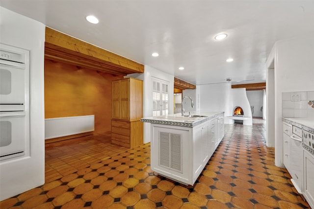 kitchen with double oven, a kitchen island with sink, sink, radiator heating unit, and white cabinetry